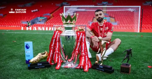 محمد صلاح يجلس على أرض ملعب أنفيلد مرتديًا قميص ليفربول، بجانبه كأس الدوري الإنجليزي الممتاز محاطًا بجوائز فردية مثل الحذاء الذهبي، لاعب الموسم، وجائزة صانع الأهداف، مع مدرجات أنفيلد الحمراء في الخلفية